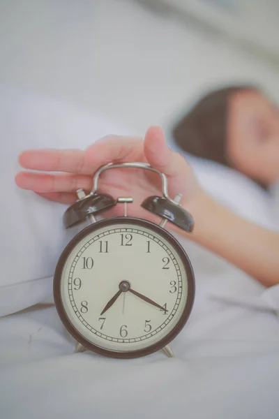 Focus on vintage alarm clock at morning time and out focus at young woman sleeping on the white bed in bedroom.