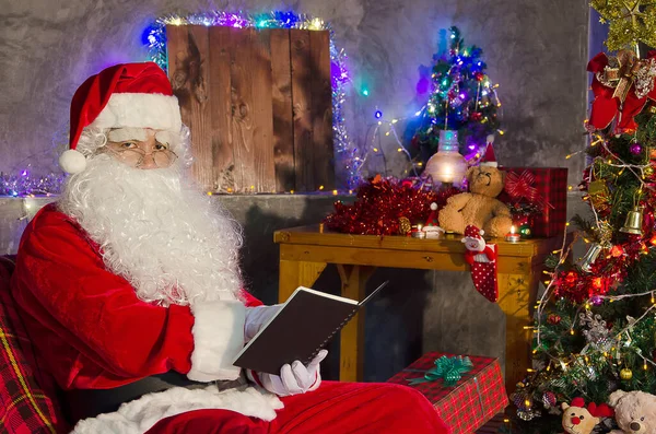 圣诞老人坐在椅子上看书 书后有一棵装饰过的圣诞树 — 图库照片