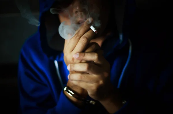 黑漆漆的照片 穿着蓝色帽带着手铐的男子在墙边吸烟 囚犯紧张地吸烟 — 图库照片