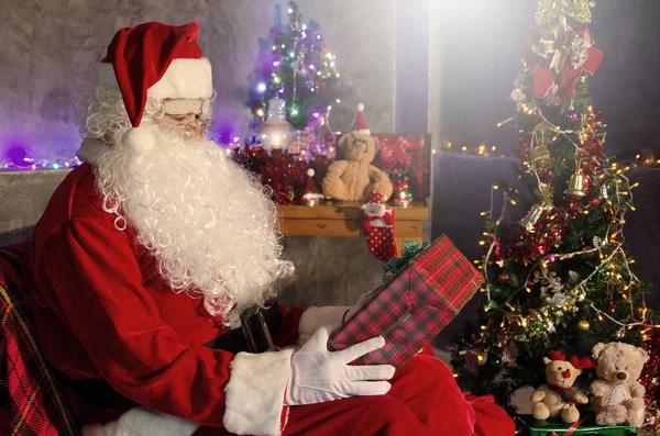 圣诞老人坐在椅子上 拿着一些礼物 背景上挂着一棵装饰过的圣诞树 — 图库照片