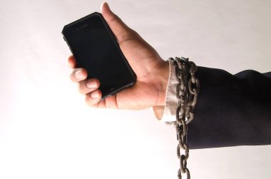 Eski çelik bir zincire sarılı, beyaz arka planda siyah bir çantada siyah bir telefon tutan takım elbiseli bir adamın bileği.
