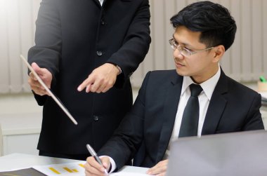 Bir iş akışı, elinde tablet tutan ayakta duran bir adamın görüntüsü ve dizüstü bilgisayarın yanında oturan ve bir şeyler yazan başka bir adama bir şeyler göstermek.