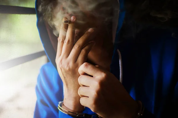 黑漆漆的照片 穿着蓝色帽带着手铐的男子在墙边吸烟 囚犯紧张地吸烟 — 图库照片