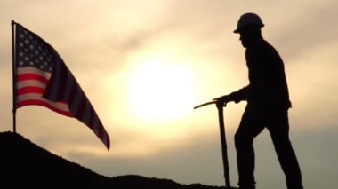 Gün batımında tepede elinde kazmayla dikilen işçilerin ve rüzgarda dalgalanan Amerikan bayrağının görüntüleri. 