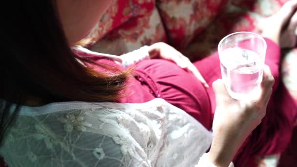 年轻的孕妇坐在沙发上喝水 — 图库视频影像
