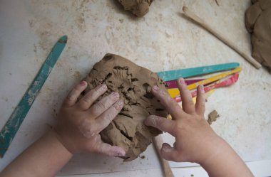 Küçük bir çocuğun elleri beyaz bir masada gri bir kil parçasıyla çalışıyor. Yukarıdan görüntüle.