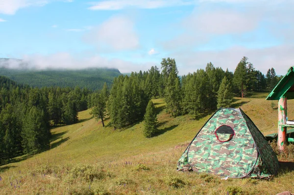 Tourist tent on top of the mountains among the forest. Wild tourism in the mountains. Camouflage tent for self-rest and hiking. Mountains, sky and white clouds. Tourism on its own