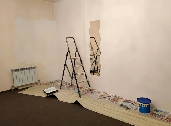 Phoography of the interior of the room during the repair. Paint the walls with paint, dirty stepladder, can of paint, floor construction. Light walls and dark floor. Mirror on the wall. Warm Artificial Light. Two walls and a corner of the room.
