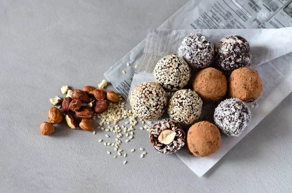 Raw vegan energy balls on the paper, gray background. Natural sweets from nuts and dried fruits in coconut, cocoa and sesame seeds, close-up. Sugar-free and gluten-free sweets, raw food desserts.