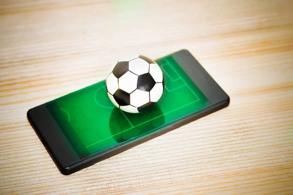Ein Spielzeugfußball auf einem Smartphone mit einem Bild eines grünen Feldes. Konzept des Fußballspiels. — Stockfoto