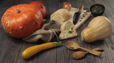 Cadılar Bayramı sebzelerinden kompozisyon. Ahşap masa arka planında turuncu ve sarı balkabakları, soğan, tohum, kabak, taze ekmek, tahta kaşıklar ve üzerinde ışık parıltısı olan bıçak var. 4k.