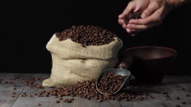 Kahve çekirdeklerinin bileşimi. Ahşap masa arkaplanı kahve çekirdekleri, kase ve kokulu kahve çekirdekleri ile süslenmiş. Eller kaseden kahve çekirdeklerini alır ve poşete döker. 4k