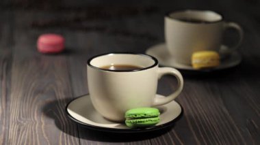 Kahve çekirdeklerinin bileşimi. Kahve çekirdekleri, renkli badem kurabiyeleri ve kahve ile süslenmiş ahşap masa arkası. El masadan bir fincan kahve alır. 4k