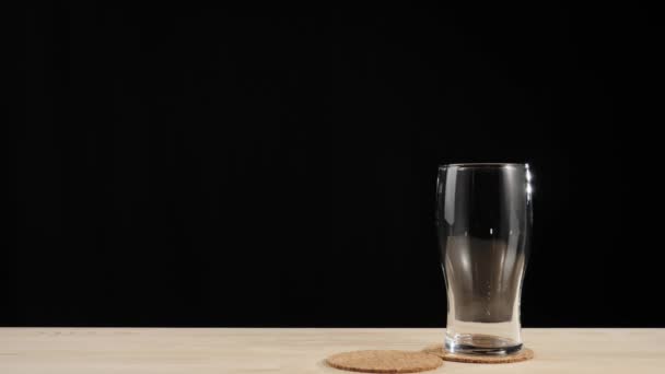 Friss sör. Kézzel tesz egy második söröspoharat egy söralátétre az első pohár mellett egy fa asztalba, fekete háttér mellett. Hideg, friss sör, vízcseppekkel. Kész az italra. Széles látószögű. 4k