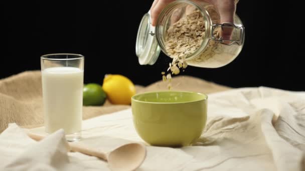 新鮮な健康食品 手はテーブルからオート麦のシリアルと瓶を取り 黒の背景に牛乳のガラスの近くの緑のボウルにオート麦のシリアルを注ぐ 健康的な朝食 きれいな食事の概念 — ストック動画