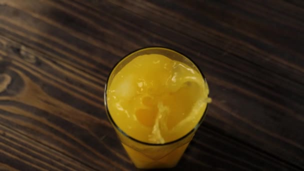 新鮮なオレンジジュースで背の高いガラスに落ちる氷のキューブのトップビューは木製のテーブルに対してスプラッシュや泡を作ります — ストック動画