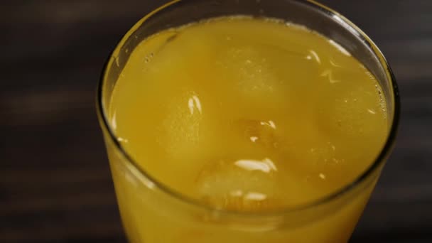 氷のキューブスピンのトップビューと木製のテーブルの上に背の高いガラスで新鮮なオレンジジュースに浮かぶ — ストック動画