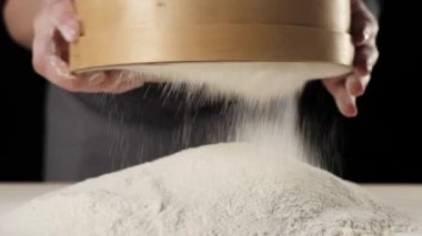 Profesyonel fırıncı elleri, masanın üzerinde siyah arka plana karşı ahşap bir süzgeçten un geçirir. Düşen beyaz parçacıklar. Hamur yoğurma ve lezzetli pizza pişirme kavramı. Kapatın. 4K