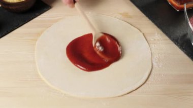 Profesyonel aşçı, pizza malzemeleriyle süslenmiş pizza tabağına ahşap bir kaşıkla kırmızı domates ezmesi sürer. Hamur yoğurma ve lezzetli pizza pişirme kavramı. Üst Manzara. 4K