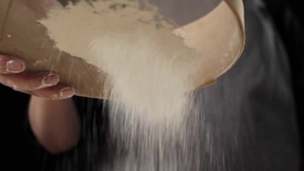Des mains de boulanger professionnel tamisent la farine à travers un tamis en bois sur la table sur fond noir. Particules blanches tombantes. Concept de pétrissage de pâte et de cuisson de délicieuses pizzas. Vue du bas. 4K 