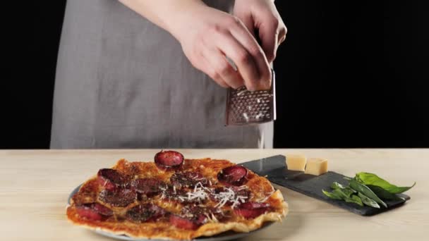 专业厨师在烤架上擦意大利芝士 烤奶酪落在比萨饼上 配上意大利腊肠和黑色背景 做美味披萨的概念中枪 — 图库视频影像