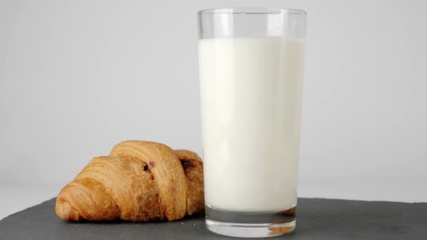 クロワッサンの近くの透明なガラスに新鮮な白い牛乳とチョコレートが白いテーブルの上の黒い板に回転します 家にいて健康的な食べ物を食べるという概念 おいしい朝食 最上階だ — ストック動画