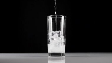 Saf taze maden suyu, siyah zemin üzerinde buz küpleri olan şeffaf bir bardağa dökülür. Evde kalıp, soda ve buzla sağlıklı yiyecekler yeme fikri. Geniş açı. Yavaş çekim