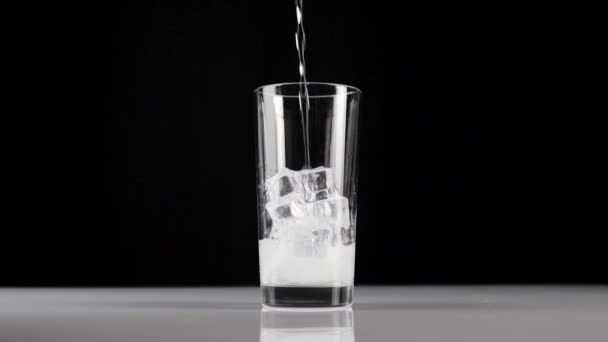 De l'eau minérale pure et fraîche est versée dans un verre transparent avec des glaçons sur fond noir. Concept de rester à la maison et manger des aliments sains avec de la soude et de la glace. Gros plan. Mouvement lent 