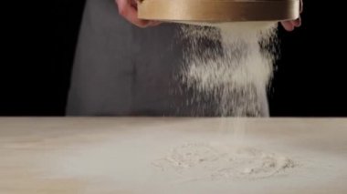 Profesyonel fırıncı elleri, masanın üzerinde siyah arka plana karşı ahşap bir süzgeçten un geçirir. Düşen beyaz parçacıklar. Hamur yoğurma ve lezzetli pizza pişirme kavramı. Yavaş çekim