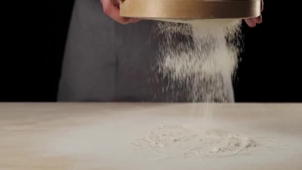 专业烘培师将面粉通过桌子上的木制筛子筛入黑色背景 坠落的白色粒子 面团揉搓和烹调美味披萨的概念 慢动作 — 图库视频影像