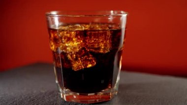 Kısa bardak kola, buz küpleri ve baloncuklar siyah masada koyu kırmızı arka planda dönüyor. Kola ya da gazoz içmek, evde rahatlamak ve eğlenmek. Kapatın. 4K