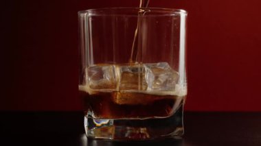 Baloncuklu lezzetli kola siyah bir masada koyu kırmızı arka planda buz küpleri olan kısa bir bardağa dökülür. Kola ya da gazoz içmek, evde rahatlamak ve eğlenmek. Kapatın. 4K