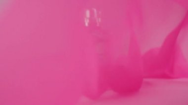 Düz yüzeyli parfüm şişesi, üzerinde cyan parfümleri ya da beyaz masa üzerinde gerekli yağlar. Pembe kumaş dalgalanır ve parfüm şişesinin etrafında dalgalanır. Aroma ve koku kavramı. Kapat.