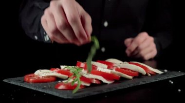 Profesyonel şef yemeğin sunumunu yapar, roka yapraklarını taze sulu domates katmanlarına serer ve siyah tahtaya mozzarella peyniri serer. Yan görüş. Kapat.