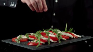 Profesyonel şef, hazır Caprese salatası ve Akdeniz bitkisi serpiyor. Bitkiler sulu domateslere, mozarella peynirine ve roka üzerine düşer. Orta atış. Kapat.