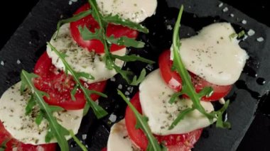 Kamera hazır Caprese salatasının üzerinde hareket ediyor. Taze sulu domatesler, mozzarella peyniri ve kara tahtada akdeniz otları ve deniz tuzu bulunan roka yaprakları. Üst Manzara. Kapat.