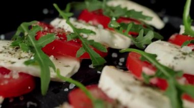 Kamera hazır Caprese salatasının üzerinde hareket ediyor. Taze sulu domatesler, mozzarella peyniri ve kara tahtada akdeniz otları ve deniz tuzu bulunan roka yaprakları. Yan görüş. Kapat.