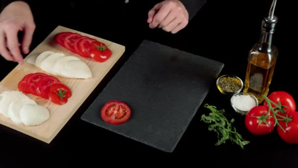 プロのシェフが料理のプレゼンテーションを行い ブラックボード上の新鮮なジューシーなトマトとモッツァレラチーズを含むカプレーゼサラダの成分をレイアウトします 撃たれた トップ表示 — ストック動画