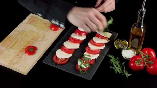 プロのシェフが料理のプレゼンテーションを行い 黒ボード上の新鮮なジューシーなトマトとモッツァレラチーズの層にアルグラの葉を配置します 撃たれた トップ表示 — ストック動画