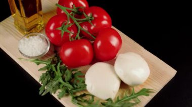 Modern Caprese salatası. Bir dalda taze sulu kırmızı domatesler, mozzarella peyniri, roka yaprakları, zeytinyağı ve deniz tuzu siyah arka planda tahtada. Kamera sola hareket ediyor. Kapat.