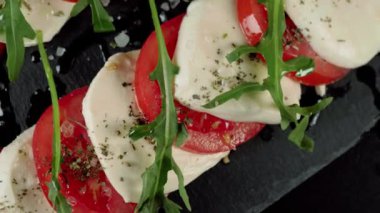 Kamera hazır Caprese salatasının üzerine çıkıyor. Taze sulu domatesler, mozzarella peyniri ve kara tahtada akdeniz otları ve deniz tuzu bulunan roka yaprakları. Yan görüş. Kapat.