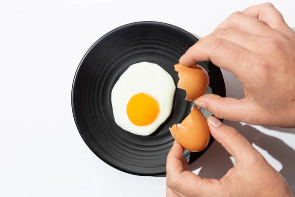 Manos agrietando Huevo frito en plato negro sobre fondo blanco. Concepto de cocina y comida . — Foto de Stock
