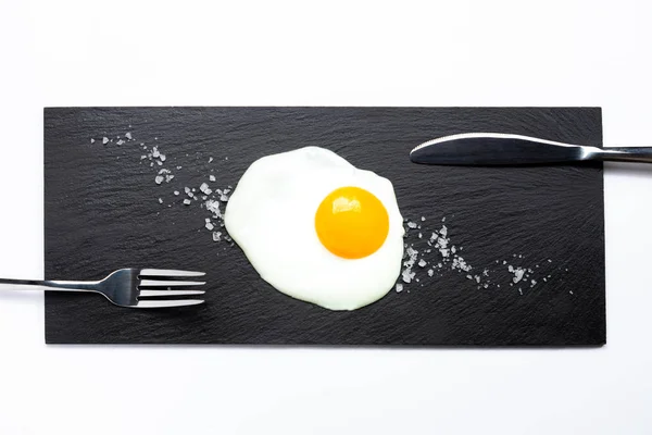 Huevo frito en plato de pizarra con cuchillo, tenedor y sal sobre fondo blanco. Concepto de cocina y comida . — Foto de Stock