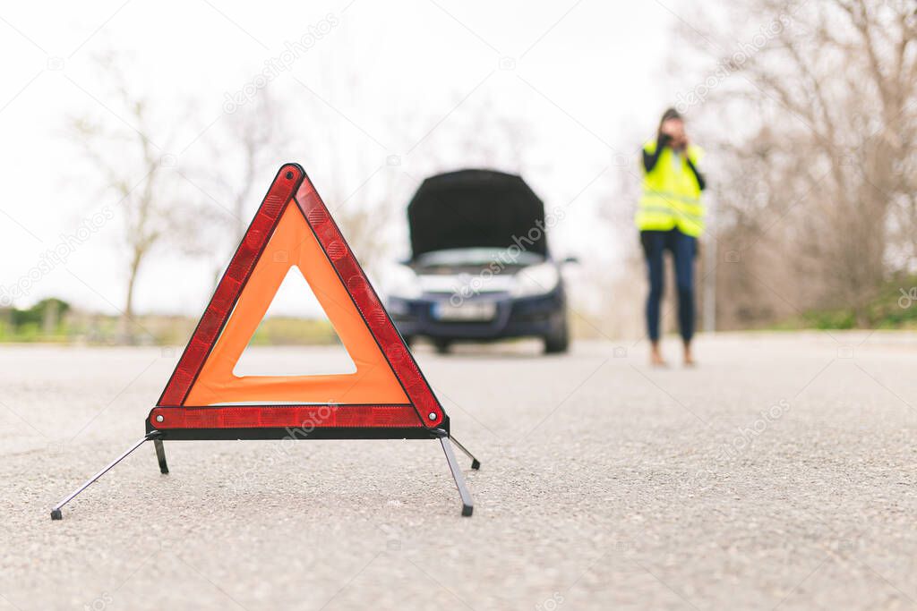 Young Caucasian woman, talking on her cell phone while her car is broken down on the road with reflective warning triangles. Selective focus. Automobile and Roadside Assistance Concept.