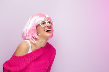 Pembe peruklu ve kazaklı, beyaz gözlüklü, fotoğraf stüdyosunda komik bir şekilde gülümseyen beyaz bir kadın. Komik anlamlı kadınlar ve renkli portre konsepti. Pembe..