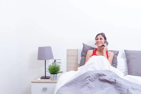 Happy young woman in bed smiling while listening on mobile phone.