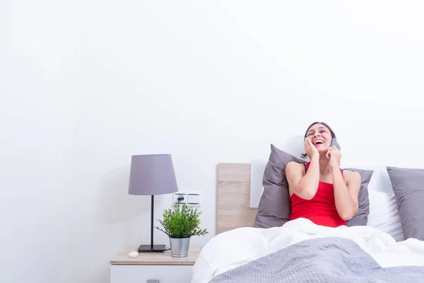 Happy young woman in bed laughing out loud while talking on the mobile phone.