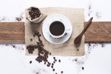 Beyaz kahve fincanı, çantadan kahve çekirdekleri dökülmüş, beyaz arka planda kahveli tahta kaşık. Üst görünüm.