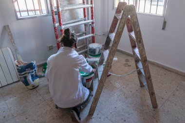 İç duvarları beyaz boyayla boyayan genç bir kadın. Boya kutuları ve boya merdiveni.
