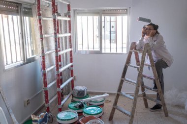İç duvarları beyaz boyayla boyayan genç bir kadın. Boya kutuları ve boya merdiveni.